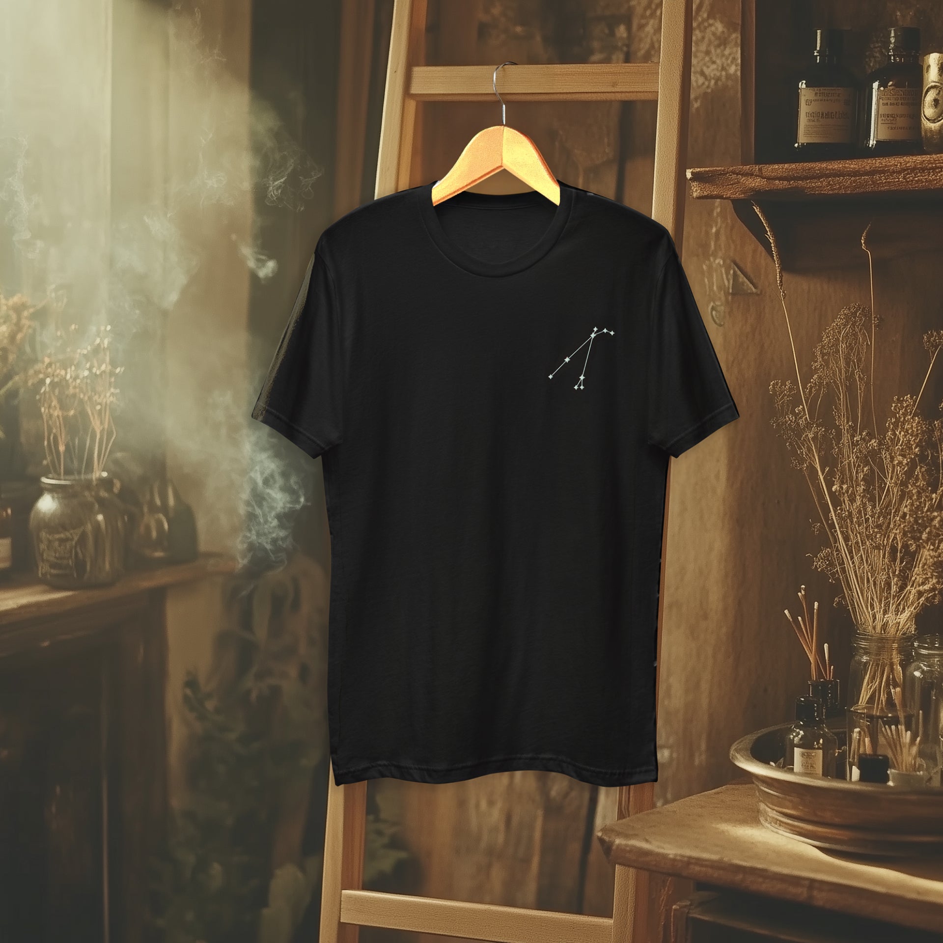 The Constellation Tee displayed on a wooden hanger in a warm, rustic setting, with smoke and dried herbs adding a mystical atmosphere.